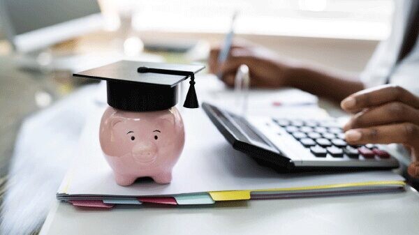 Piggy bank with a graduation hat on a desk with a person using a calculator.