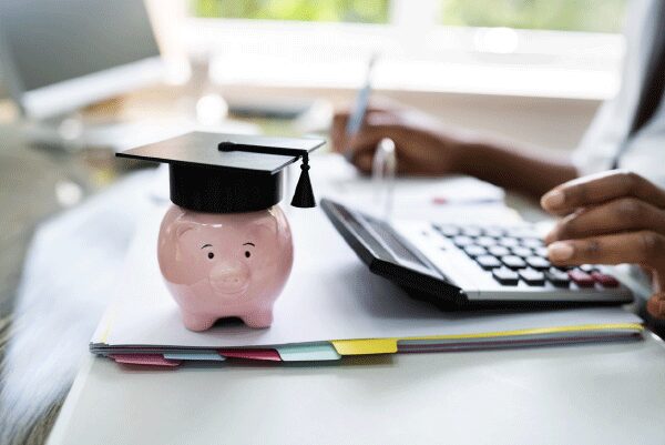Piggy bank with a graduation hat on a desk with a person using a calculator.