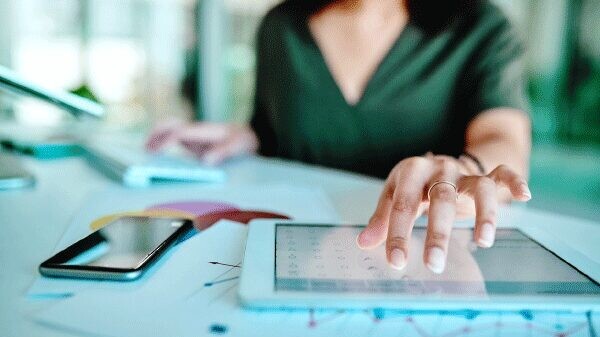 Woman using tablet with cell phone and printed charts on the table.