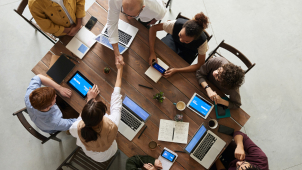 Arial view of 8 workers with laptops seated around a wooden conference table.