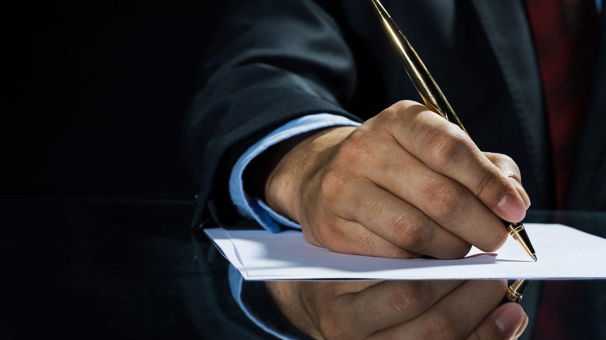 Person clothed in a business suit, signing a document with a gold pen.