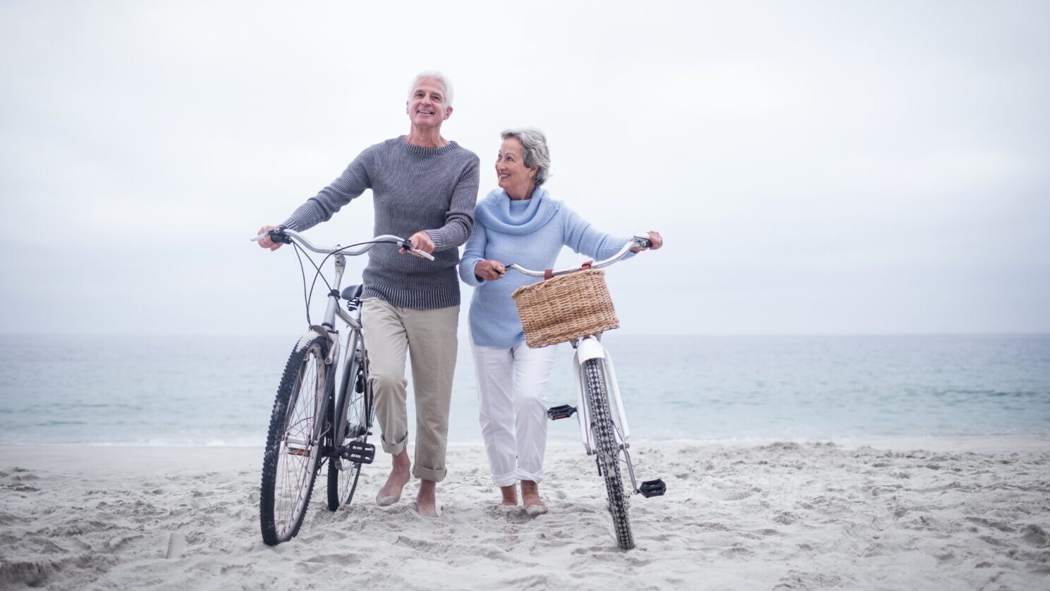 Older man and woman on the beach pushing bicycles.