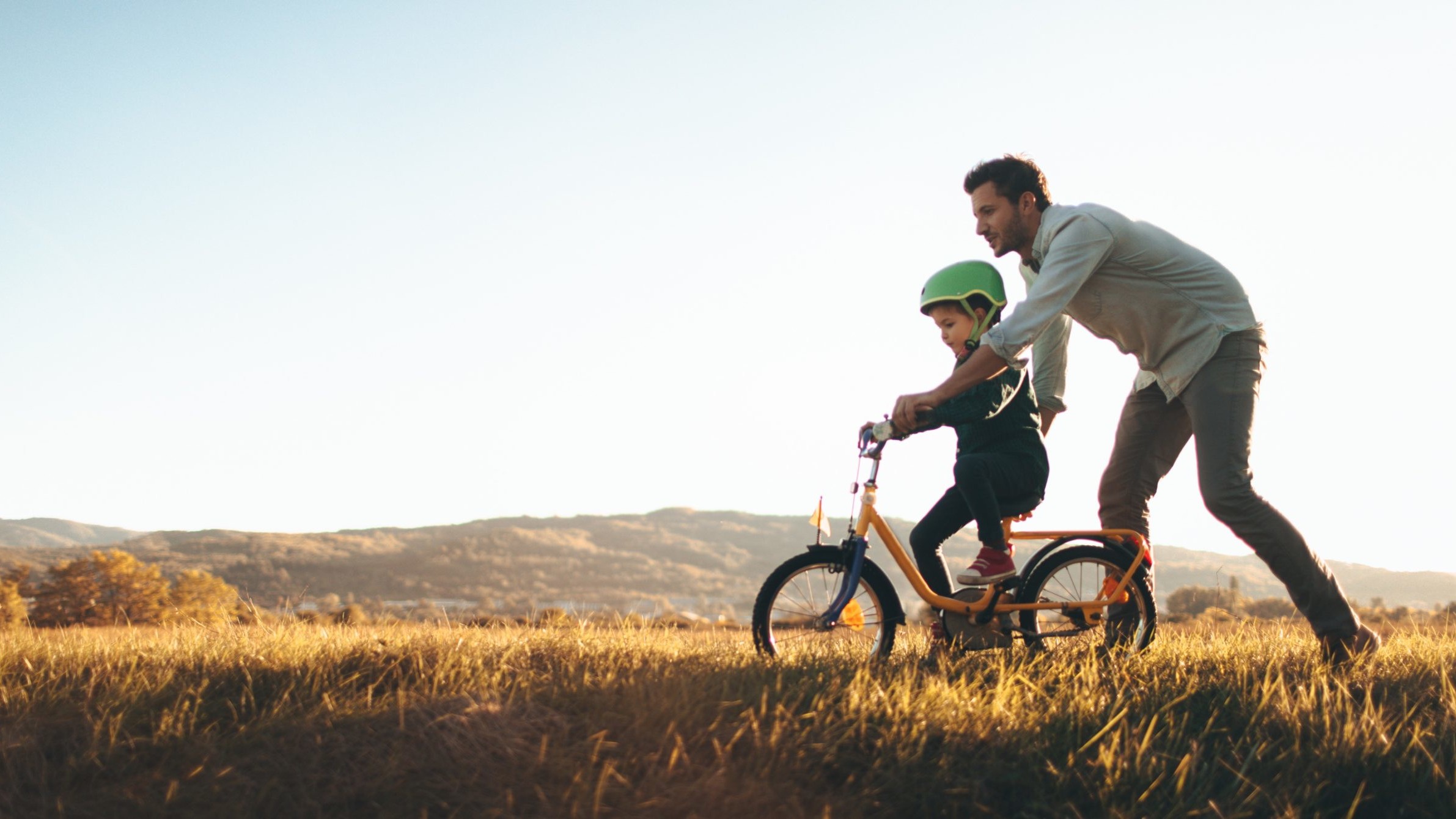 A small child on a bike, being helped by a man.