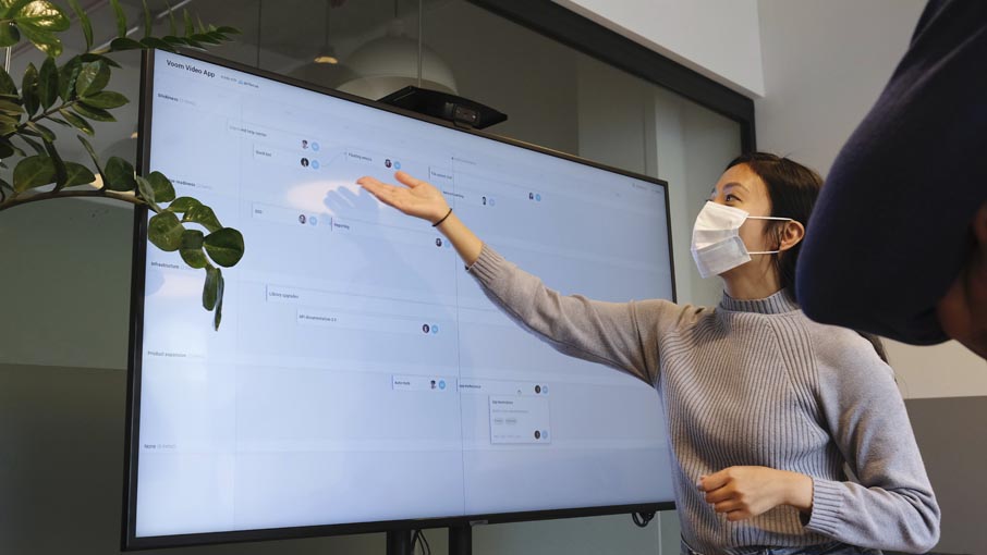 A masked woman, making a presentation in front of a large screen.