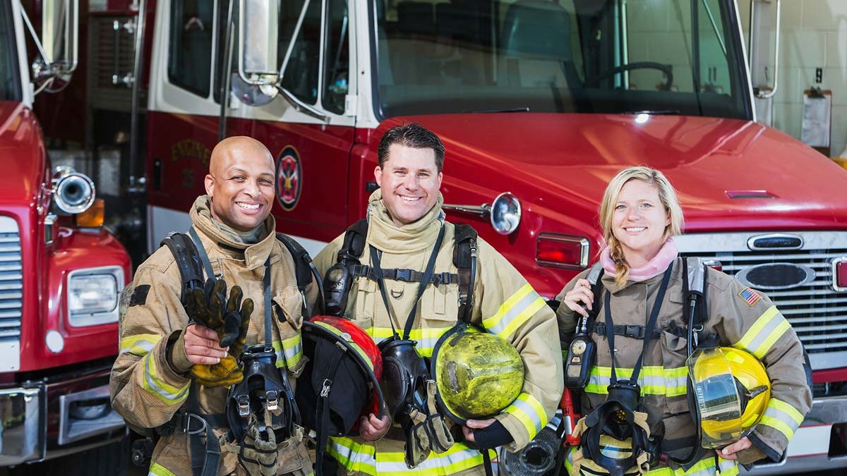 Three firefighters, standing in front of firetrucks.