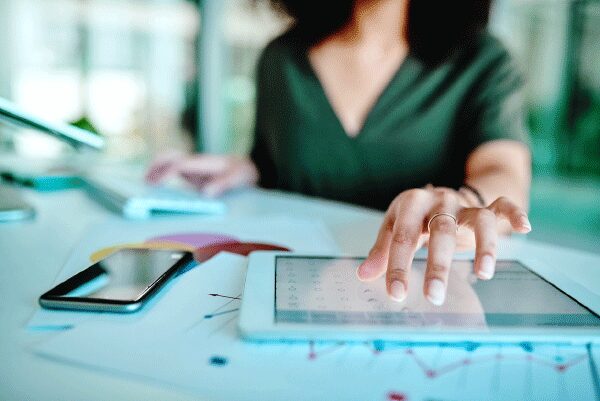 Woman using tablet with cell phone and printed charts on the table.