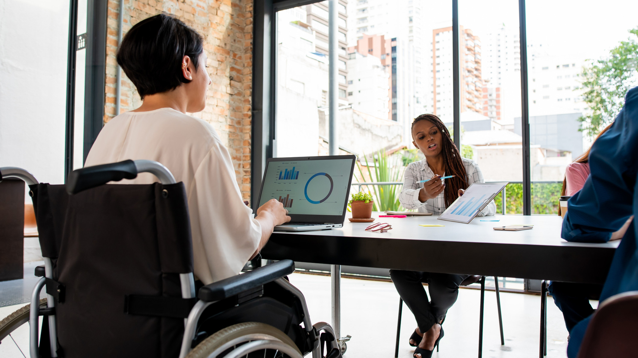 A woman in a wheelchair works on a laptop, while across the table, a Black woman shows her a chart.