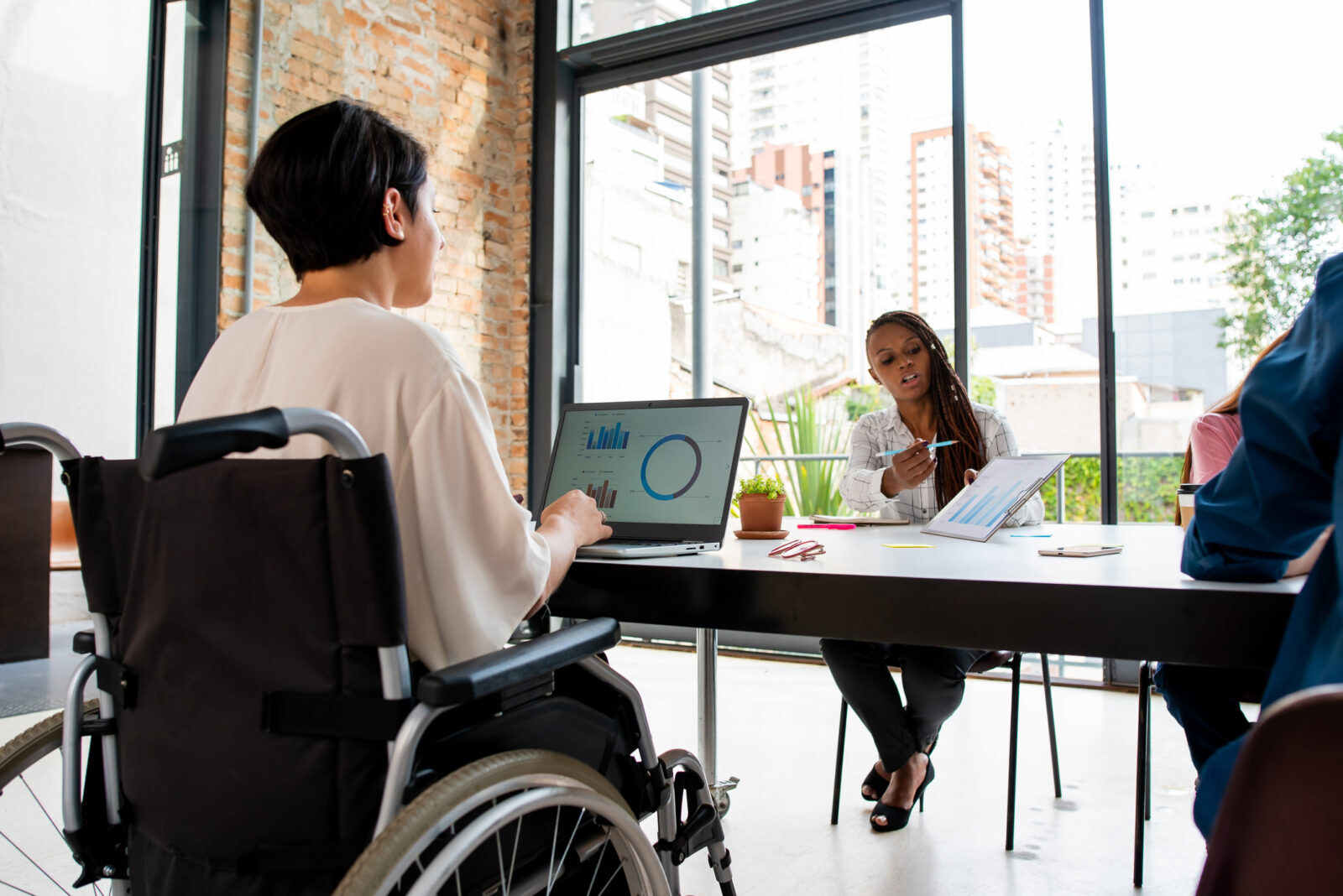 A woman in a wheelchair works on a laptop, while across the table, a Black woman shows her a chart.