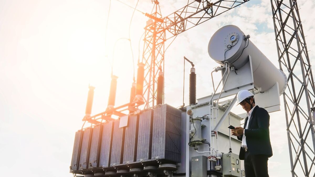 A low angle image of a businessman wearing a black suit, standing looking at a large power transformer with blue sky to be background, Concept about business people who want to invest in energy