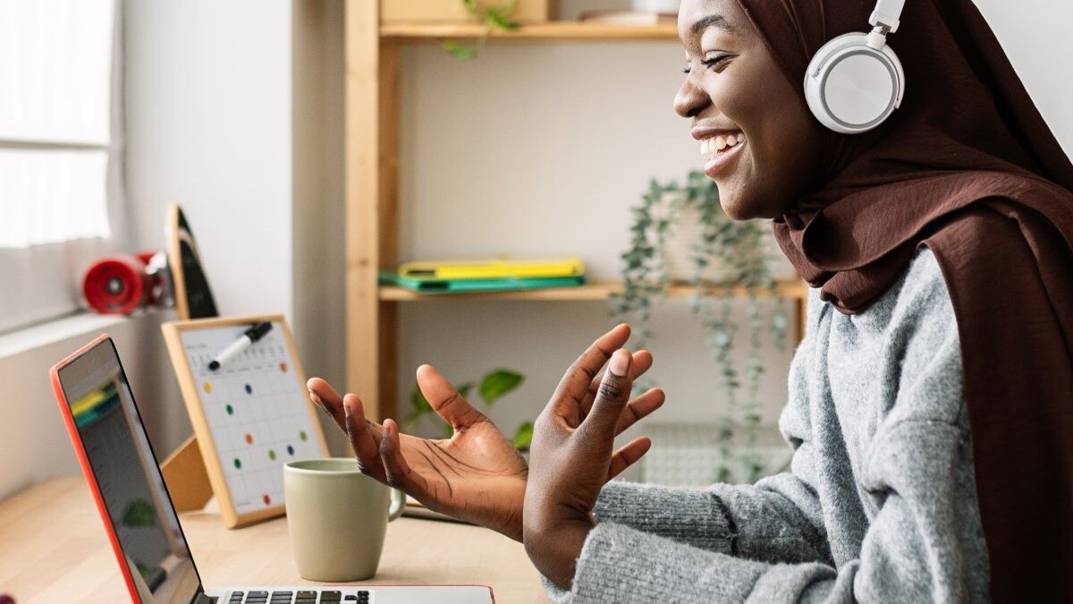 Happy female student in muslim headscarf having a video call on laptop at home. Millennial female enjoying online virtual meeting conference with friend