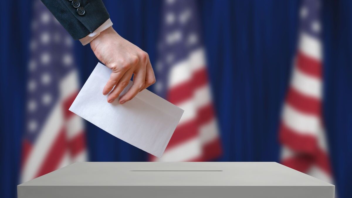 Hand submitting a vote into an election box, United States flags shown in background