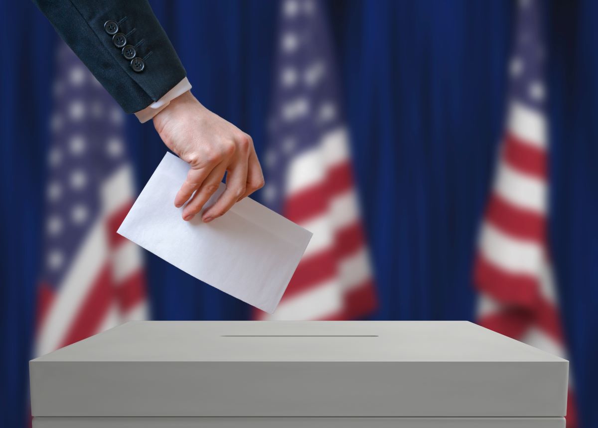 Hand submitting a vote into an election box, United States flags shown in background