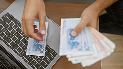 Young adult sitting on his living room sofa counting yuan banknotes managing his wealth via laptop at his cozy home