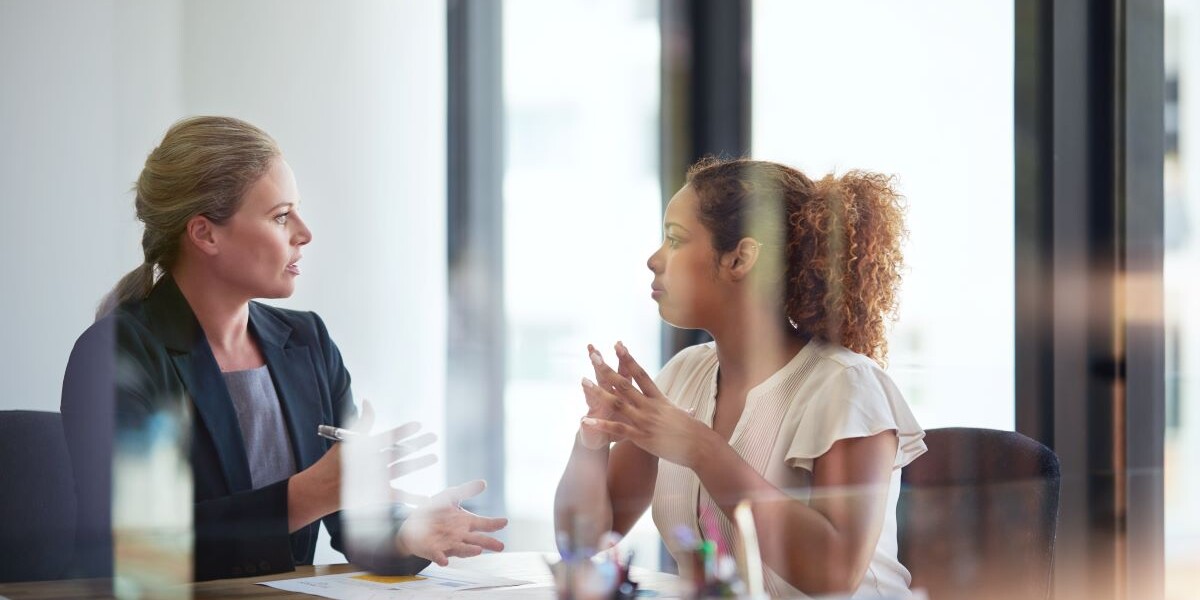 two women speaking at a conference table in a meeting, perspective through glass doors.