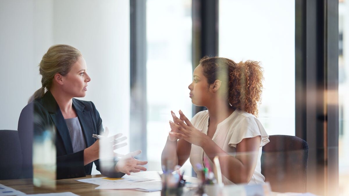 two women speaking at a conference table in a meeting, perspective through glass doors.