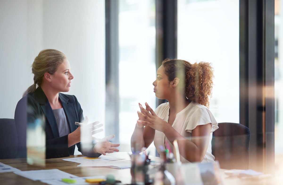 two women speaking at a conference table in a meeting, perspective through glass doors.