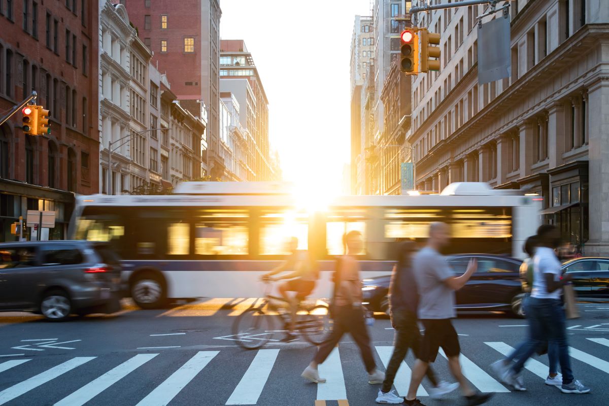 People, cars, bikes and buses traveling through a busy intersection on 5th Avenue and 23rd Street in New York City with shining sunlight background