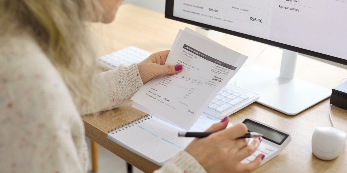 Senior woman calculates electricity bills with calculator. Close view at the desk shows paperwork as she tracks expenses, updates budget, and schedules payment.