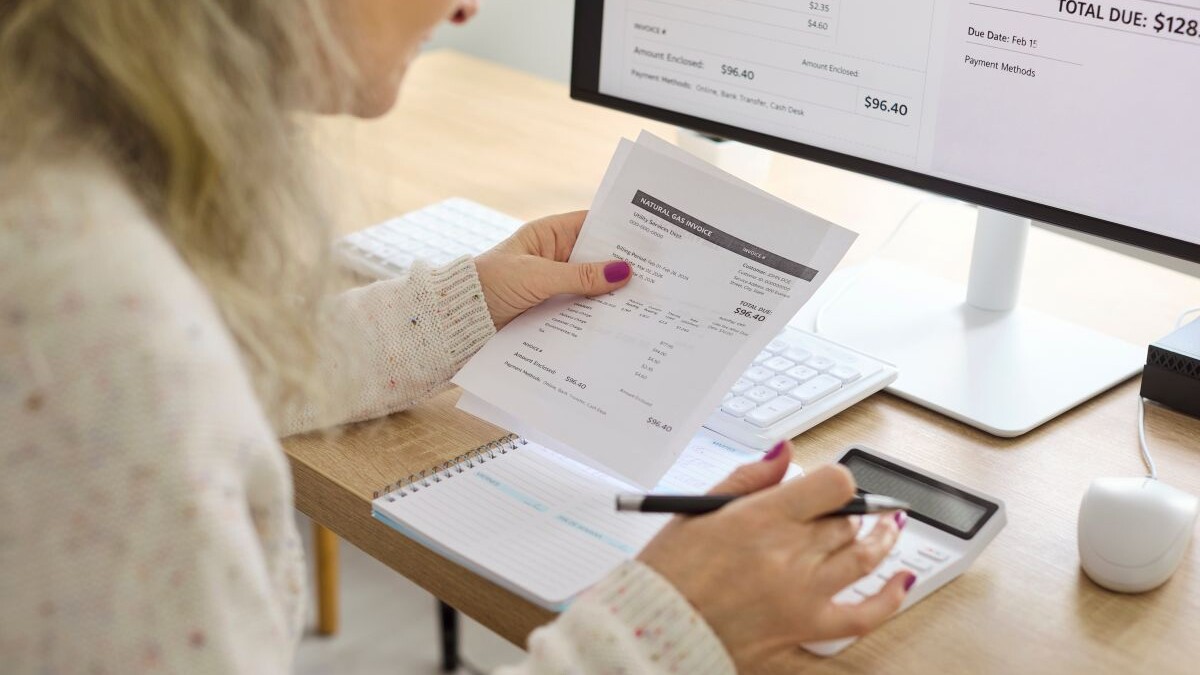 Senior woman calculates electricity bills with calculator. Close view at the desk shows paperwork as she tracks expenses, updates budget, and schedules payment.