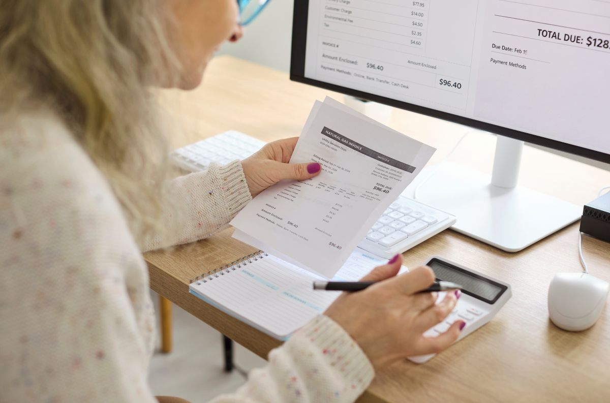 Senior woman calculates electricity bills with calculator. Close view at the desk shows paperwork as she tracks expenses, updates budget, and schedules payment.